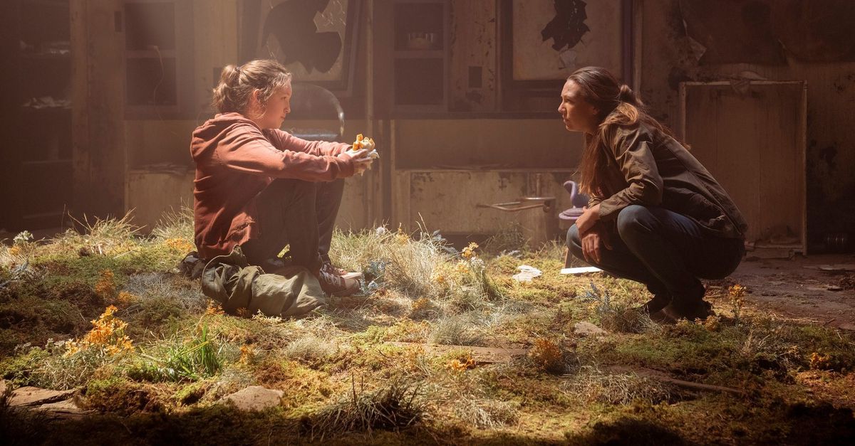 Bella Ramsey as Ellie and Anna Torv as Tess in HBO's 'The Last of Us.' Ellie is sitting and Tess is crouching on overgrown grass inside an abandoned building as they talk. Ellie is a white teenage girl with her brown hair in a ponytail, a red hoodie, and jeans. Her backpack is at her feet, and she's eating a sandwich. Across from her is Tess, a white woman with long brown hair, wearing a brown buttondown and jeans.