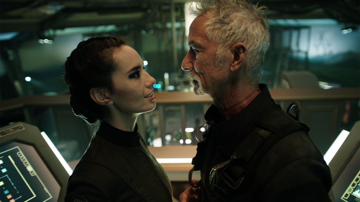 Cara Gee as Camina Drummer (left) and David Straithairn as Klaes Ashford (right) on 'The Expanse.' Drummer is a light-skinned Native woman with her dark hair braided into a tight bun. She's wearing a high-collared black shirt. Ashford is an older white man with short, white hair and a white handlebar mustache. He's wearing a high-collared black shirt and a harness. They are standing facing each other at the control panels on a space ship.