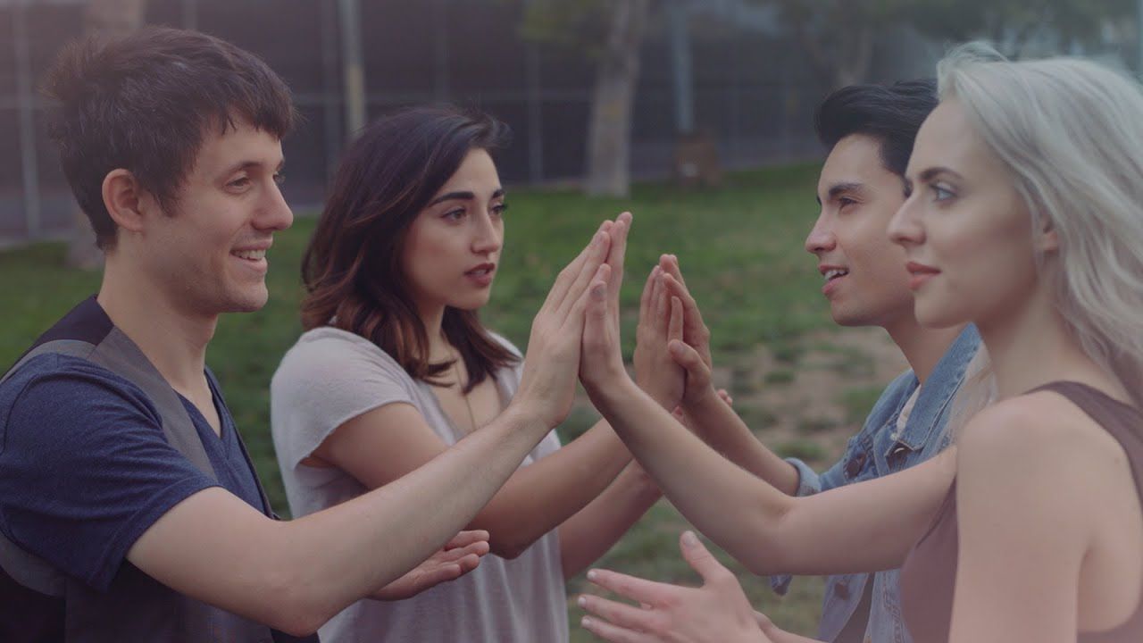 Kurt Hugo Schneider, Sam Tsui, Madilyn Bailey, and Alex G in their "Send My Love (to Your New Lover)' video (2016). They are sitting in a park and seated in a square so that they can play patty cake with someone in front of them and to their side. 