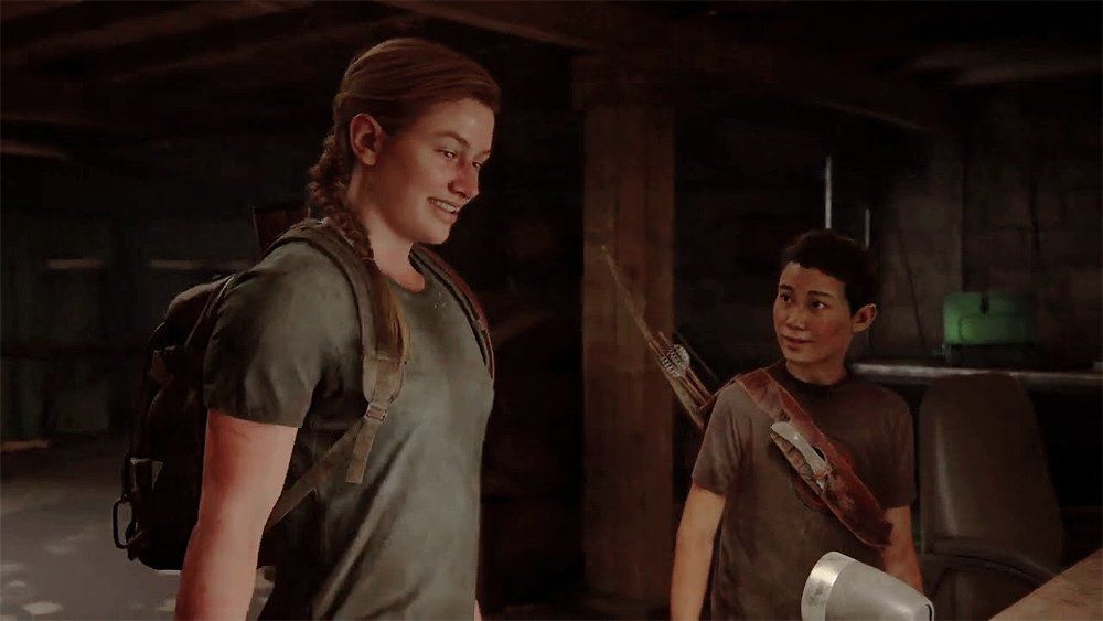 Abby (left) and Lev (right) in a scene from the video game 'The Last of Us, Part II' (2020). Abby is a white teenage girl with a muscular build and her blonde hair pulled back into a braid. She's wearing a grey t-shirt and a brown backpack. She's smiling as she looks down at something. Lev is an Asian trans boy who is shorter than Abby. He has close-cropped black hair and is wearing a brownish t-shirt with a quiver of arrows and a bow strapped to his back. He's looking at Abby. They are in a basement standing at a table with a radio on it. 