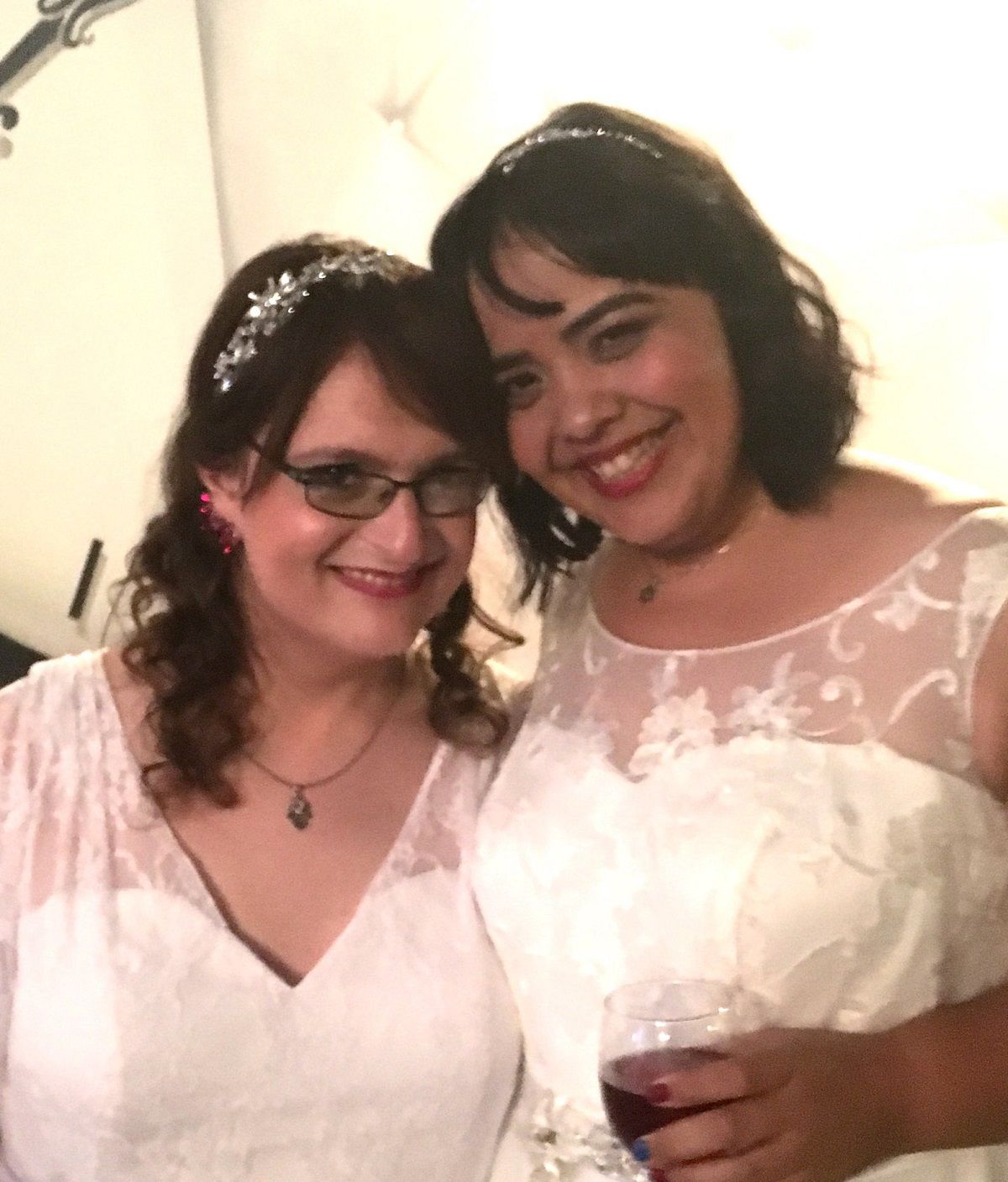 Ada Douglass (left) and Teresa Jusino (right) at their wedding. Teresa has her arm around Ada and is holding her close as they both smile for the camera. Ada is a white Jewish woman with shoulder-length brown hair with side bangs and a glittery headband in her hair. She's wearing red earrings, a hamsa necklace, and a wedding dress with a v-neckline and sheer lace around the shoulders. Teresa is a brown skinned latina with chin length black hair and bangs. She's wearing red lipstick and a star of david necklace. Her wedding dress is sleeveless, and there's sheer lace at the top just above the bodice. Teresa is holding a glass of red wine.