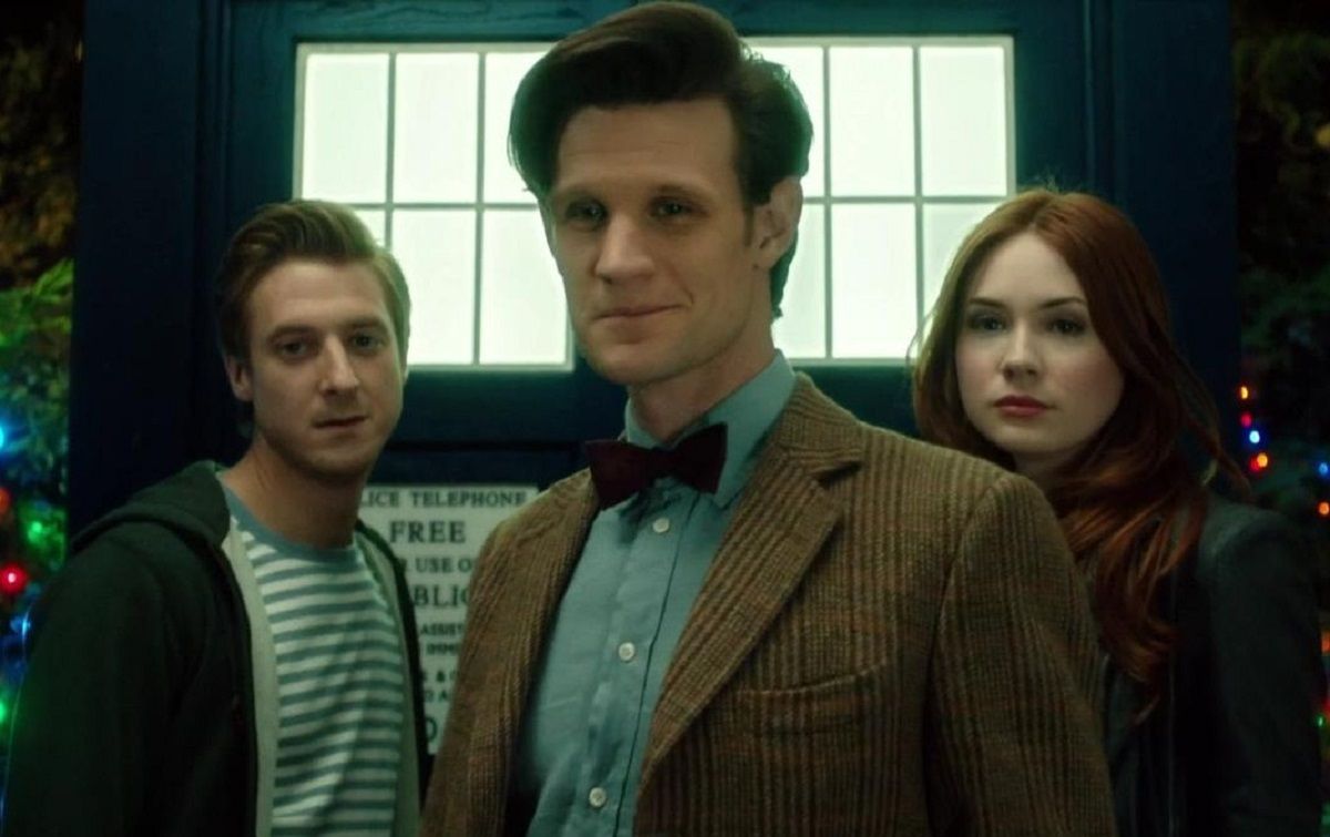Rory (Arthur Darvill), The Doctor (Matt Smith) and Amy (Karen Gillan) in a scene from 'Doctor Who.' They are standing in front of the TARDIS (a blue police phone box). They are all white. Rory is a man with medium length brown hair wearing a blue and white striped shirt and a black hoodie. The Doctor has thick dark hair in a pompadour and wears a tweed blazer over a blue buttondown with a bow tie. Amy has long red hair and wears a black jacket.
