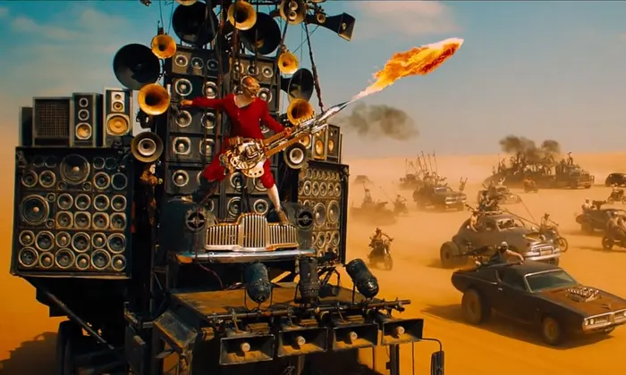 The Doof Warrior in 'Mad Max: Fury Road' (2015). He's a man dressed in red long johns and wearing a helmet that covers his eyes and playing a guitar that blows fire as he's tied to a ridiculous amount of enormous speakers on top of a car. In the background are other tricked out cars driving through the desert. 