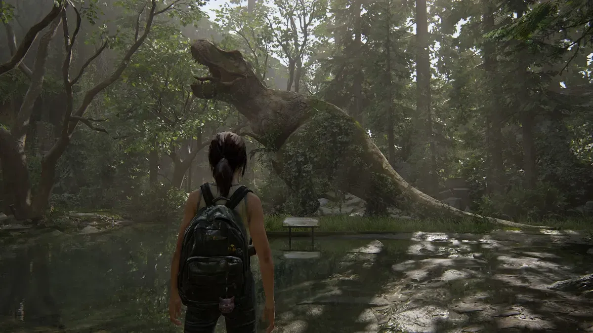 Ellie has her back turned to the camera as she comes upon the large statue of a tyrannosaurus rex in an overgrown wooded area. She has brown hair in a ponytail and wears a light colored tank top, jeans, and a backpack.