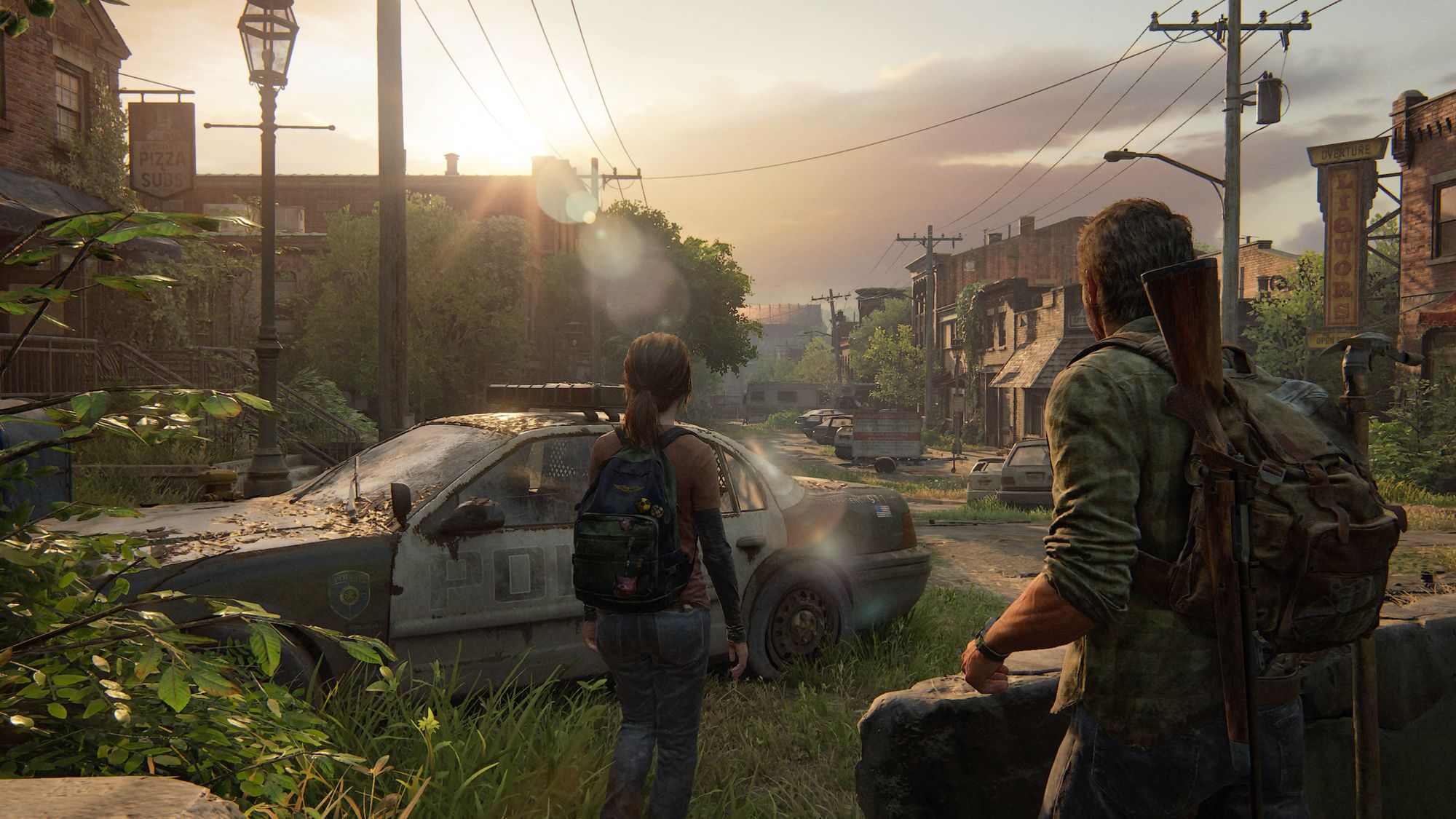 Ellie and Joel in a scene from the video game, 'The Last of Us' (2013). We see them from the back. Joel is in the foreground. He has short brown hair and is wearing a green plaid button down and a backpack with rifles and a piece of pipe affixed to it. Ellie walks ahead of him, her shoulder-length brown hair in a ponytail. She's wearing a red t-shirt over a long-sleeved black shirt, jeans, and a green backpack. They are making their way around abandoned cars as they walk down an overgrown, abandoned small town street.