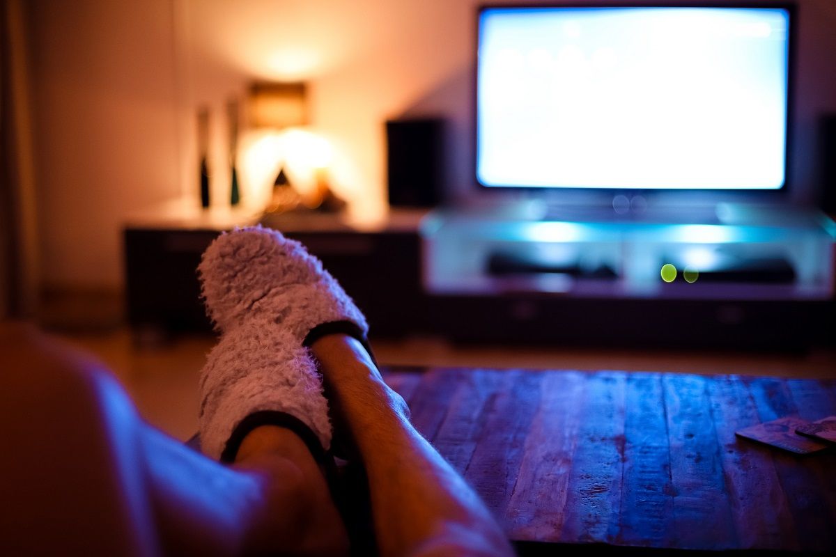 Image of a person wearing fuzzy slippers watching TV. In focus in the foreground is a person's legs up on a table in the fuzzy slippers. Out-of-focus behind that is a home entertainment system with a TV that is on, but the screen is just white light. 