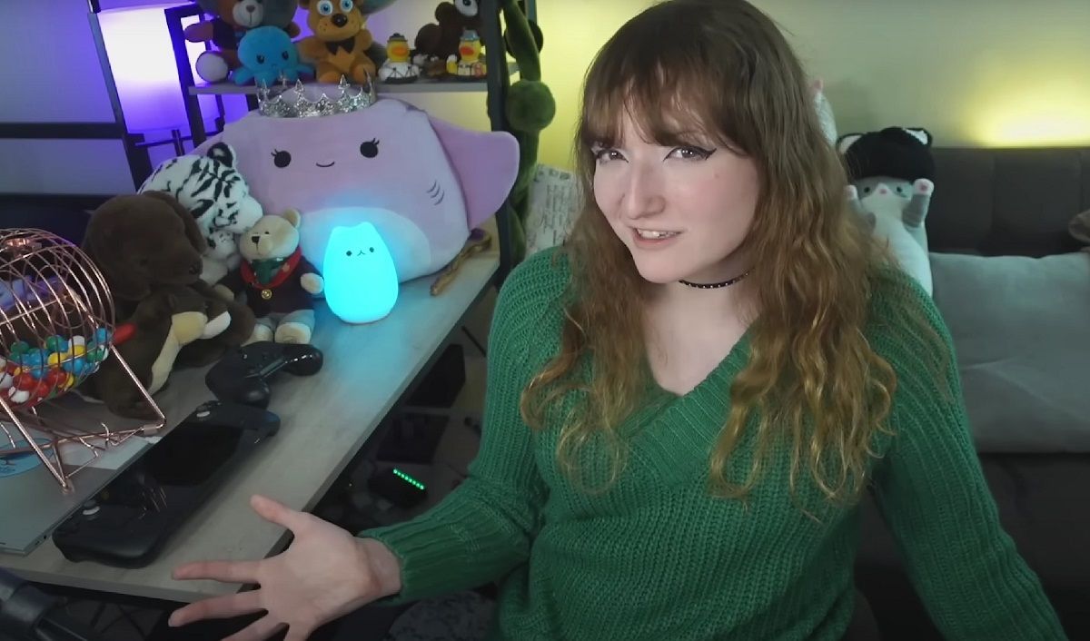 YouTuber Cara aka "heyitscara." She is a young white woman with long strawberry blonde hair and bangs. She's wearing a long-sleeved green v-neck sweater and a black choker around her neck. She's sitting next to a table with game controllers and lots of plushies on it. Behind her is a couch with yet more plushies.