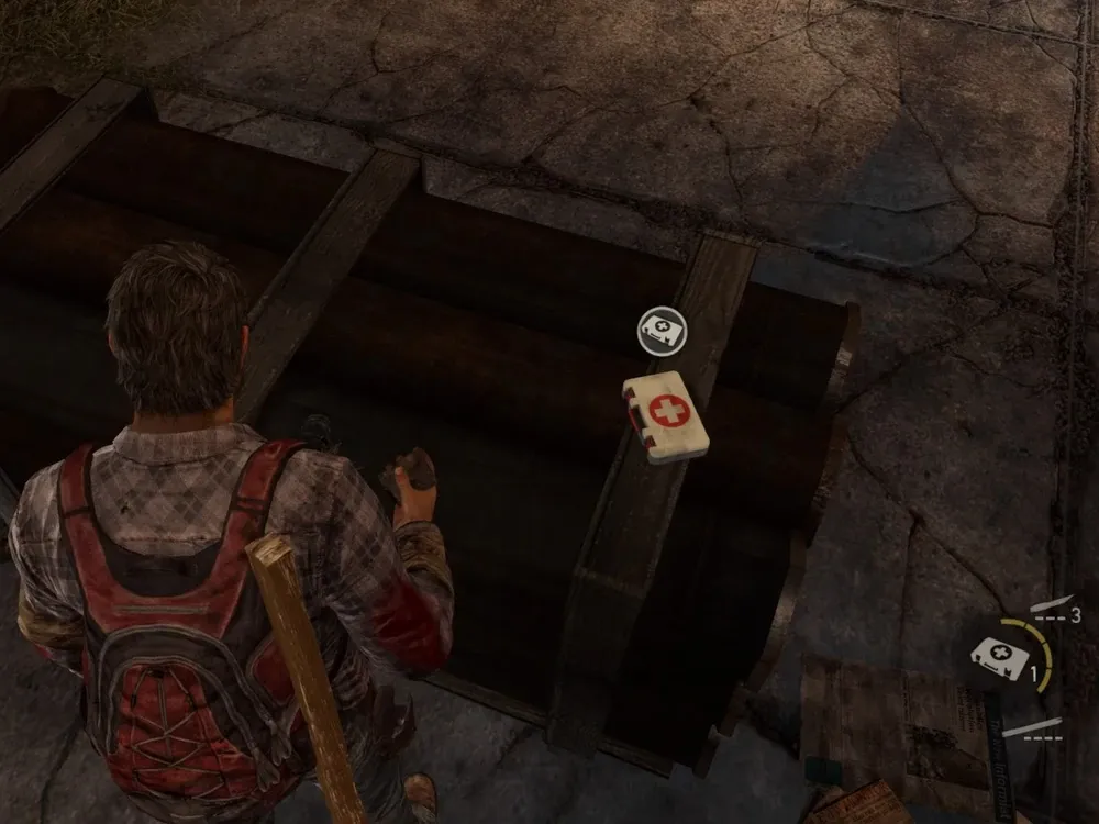 Joel in a scene from the video game 'The Last of Us' (2013). We see Joel from behind and above. The view is during gameplay. Joel is a white man with short, brown hair. He's wearing a brown plaid buttondown shirt and a red and black backpack with a plank of wood affixed to it. He stands in a dark room over pipes holding a rag in his hand and there is a health kit (a white plastic box with a white cross in a red circle on it). There is an indicator of how many health kits the player has (1) in the lower right hand corner of the image. The player also has 3 shivs and has full use of the plank of wood.