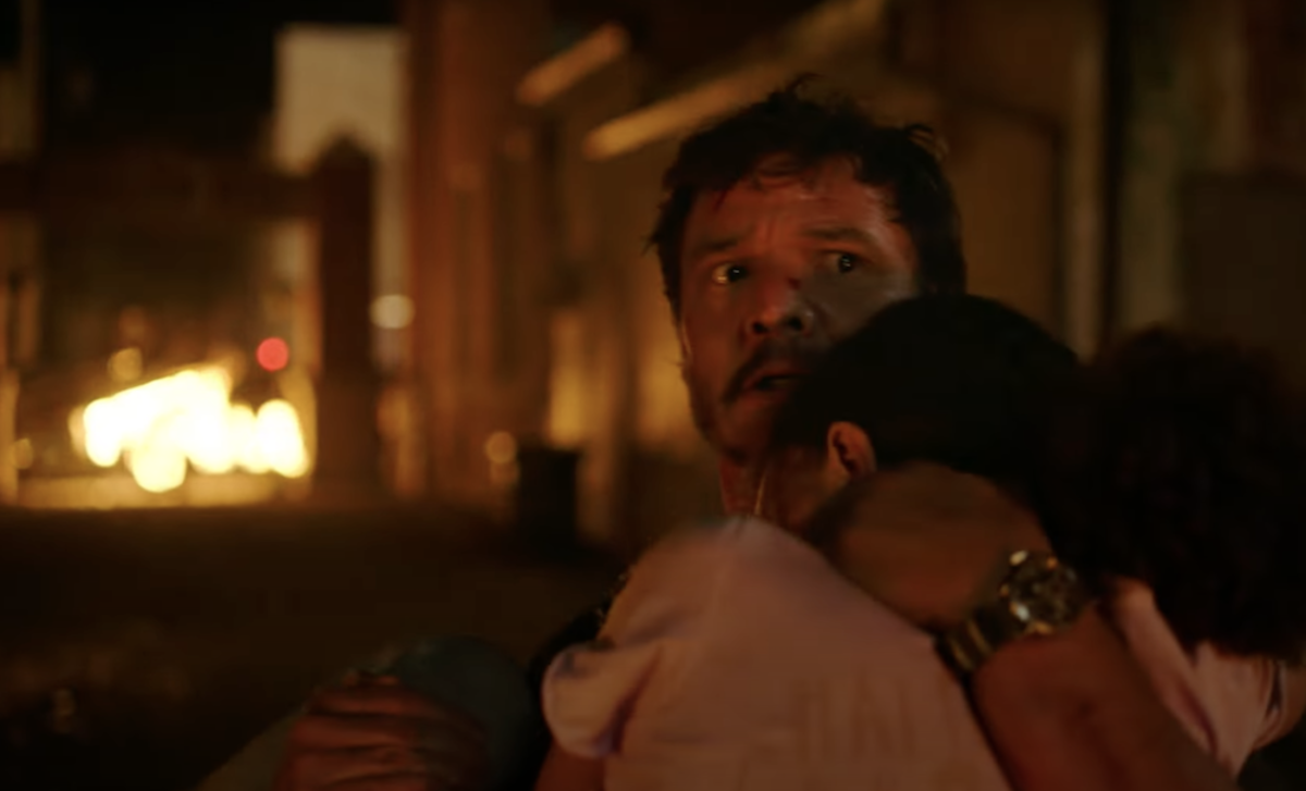 Joel (Pedro Pascal) carries Sarah (Nico Parker) down the street at night as he worriedly looks over his shoulder. There is something on fire in the background.