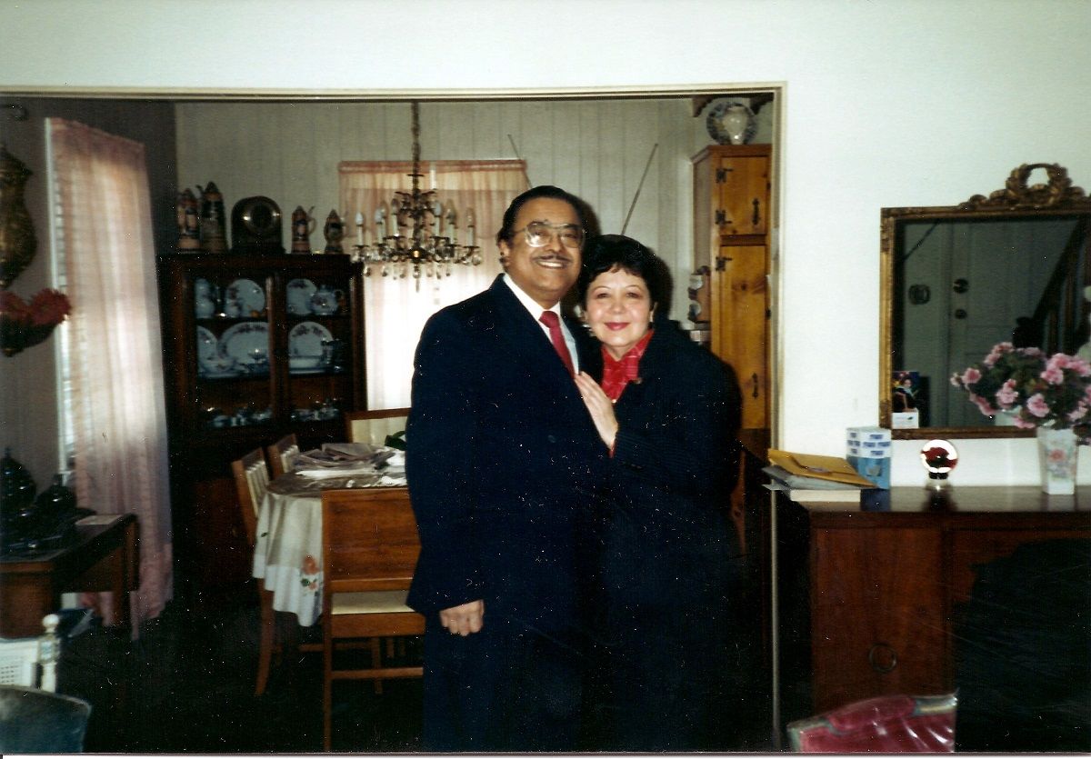 Ramon Jusino Jr (left) and Mariana Hernandez Jusino (right). Ramon is a dark-skinned Puerto Rican man with short black hair and a mustache. He's wearing glasses and a dark suit with a white buttondown and a red tie. He has his arm around Mariana, who is shorter than him. She is a white Puerto Rican with short black hair and wearing red lipstick, and a red blouse under a black skirt suit. They are standing in a living room with the dining room table in the background.