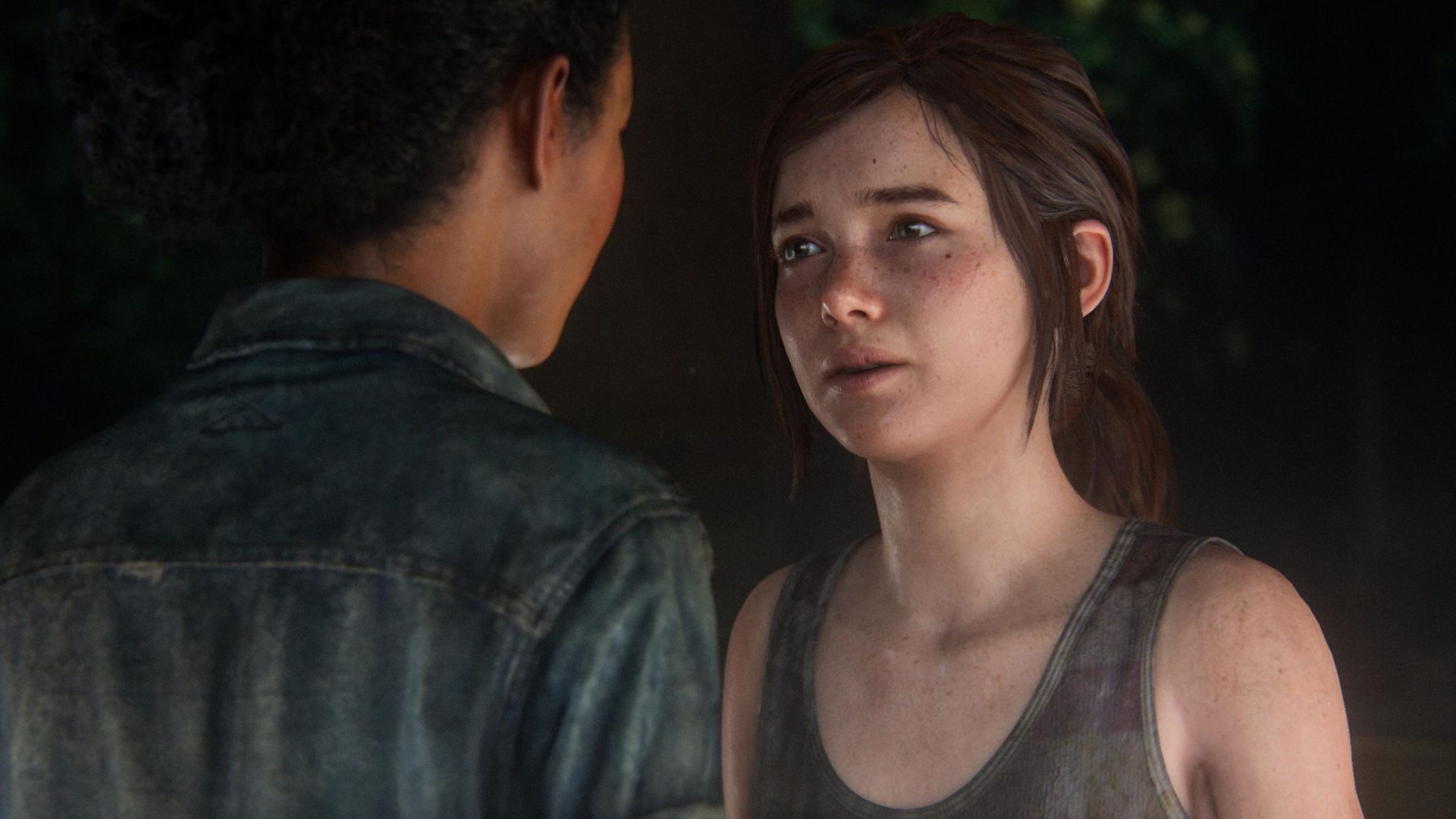 Riley (left) and Ellie (right) in a scene from the "Left Behind" DLC (2014). Riley, a Black teenage girl, has her back to the camera. She's wearing a denim jacket and has her black hair pulled back into a bun. Ellie is a white teenage girl who is looking seriously into Riley's eyes. Her long brown hair is pulled back and she's wearing a tank top.