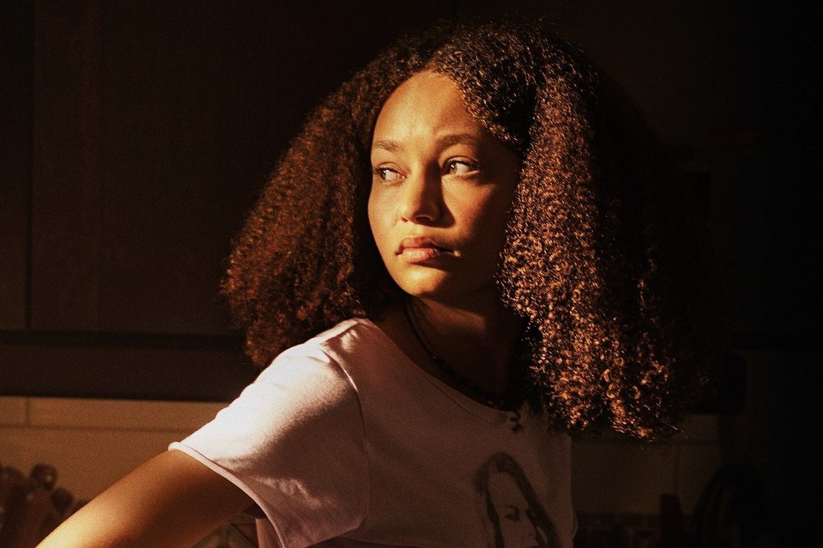 Nico Parker as Sarah. She's a Black teenage girl with shoulder length curly hair and wearing a pink t-shirt. She's looking over her shoulder.