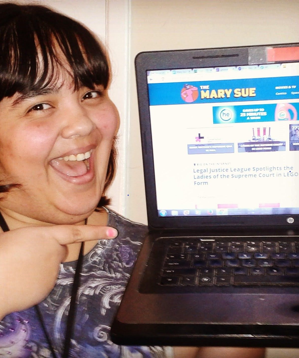 Teresa Jusino showing off her first post at The Mary Sue (2015). She is a fat, olive-skinned latina with chin-length black hair and bangs. She's wearing a purple paisley shirt with a lanyard around her neck as she smiles wide and points at the laptop she's holding in her other hand. On the screen is her first post titled "legal justice league spotlights the ladies of the Supreme Court in lego form"