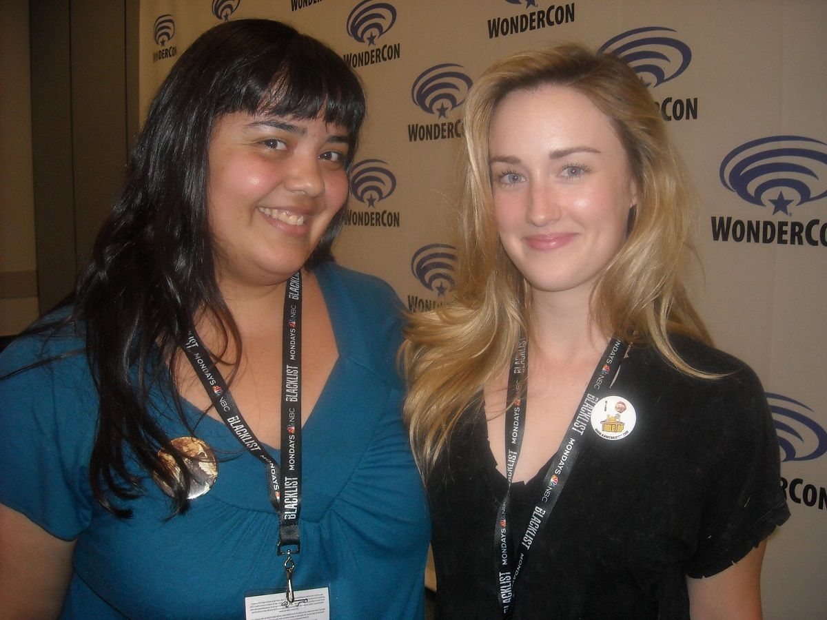 Teresa Jusino (left) and Ashley Johnson (right) at WonderCon (2017). Teresa is a brown skinned latina with long dark hair and bangs. She's wearing a blue-green v-necked shirt with short sleeves, a lanyard around her neck with a WonderCon badge, and a Doctor Who button on her shirt with the Third Doctor and Sarah Jane on it. Ashley is a white woman with long strawberry blonde hair. She's wearing a black v-necked shirt with short sleeves and a badge lanyard. She's wearing a white button. Both are smiling for the photo in front of a WonderCon step and repeat. 