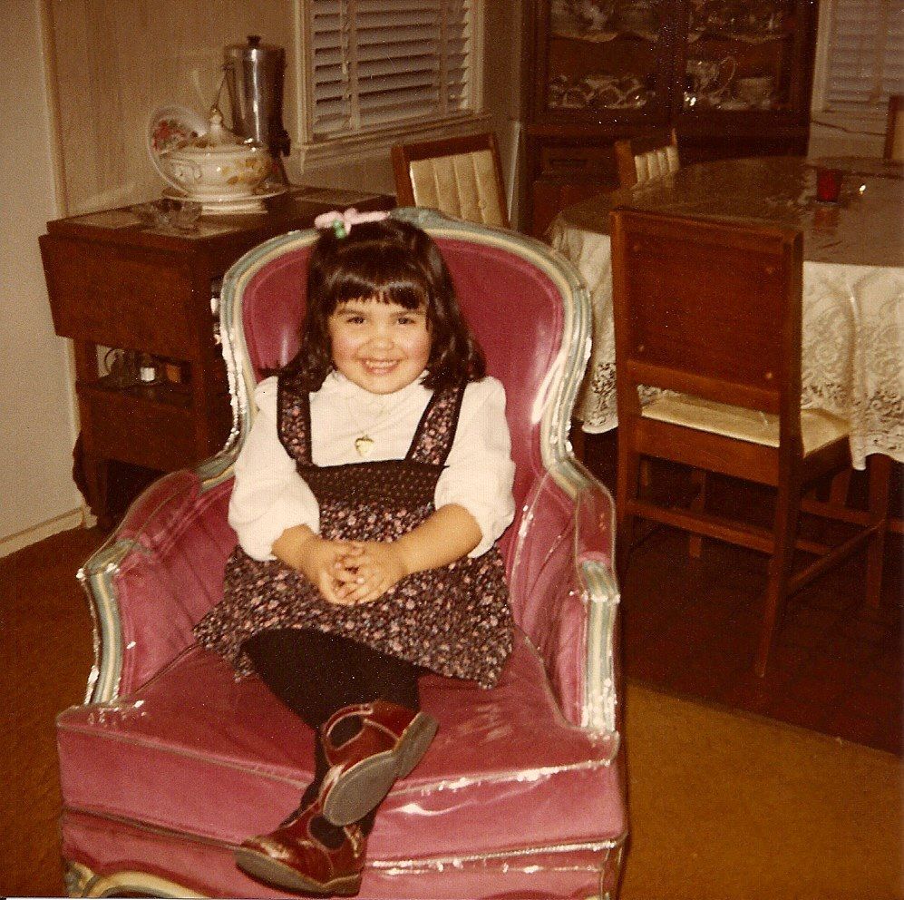 Teresa Jusino at five years old at her grandmother's house (circa 1984). Teresa is an olive complexioned latina sitting in a pink armchair with a plastic cover. She has shoulder-length dark hair with bangs and some of it tied up into a small ponytail on the side of her head. She's wearing a long-sleeved, collared white shirt under a black velvet floral print dress with thick shoulder straps. She has a gold heart-shaped locket around her neck and wears black tights and red mary jane shoes. Her legs are crossed at the ankles and her hands are folded together as she smiles for the picture. Behind her is a view of the dining room table. 