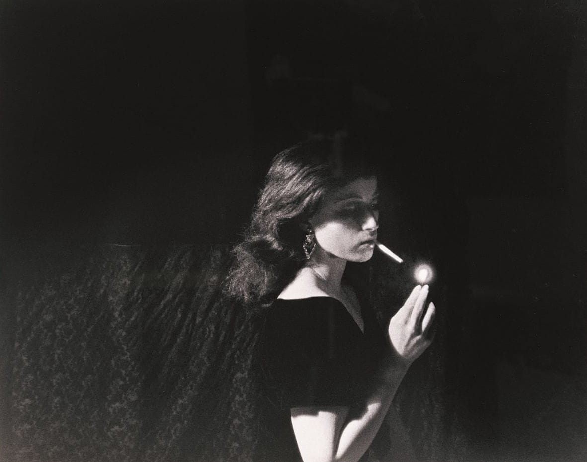 Black and white image of a white woman with shoulder length, wavy dark hair standing in an empty black room wearing dangling earrings and a black dress lighting a cigarette in her mouth.