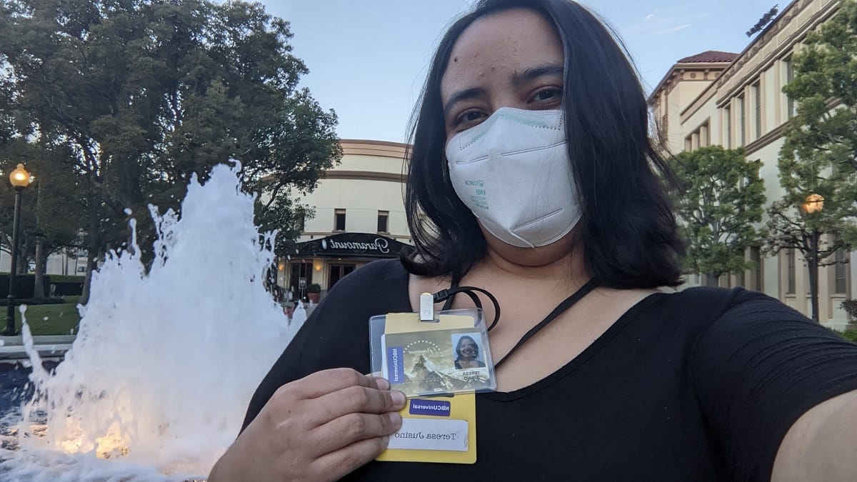 Image of a brown Latina with shoulder-length dark hair in a black t-shirt standing in front of a fountain holding up a Paramount name badge.