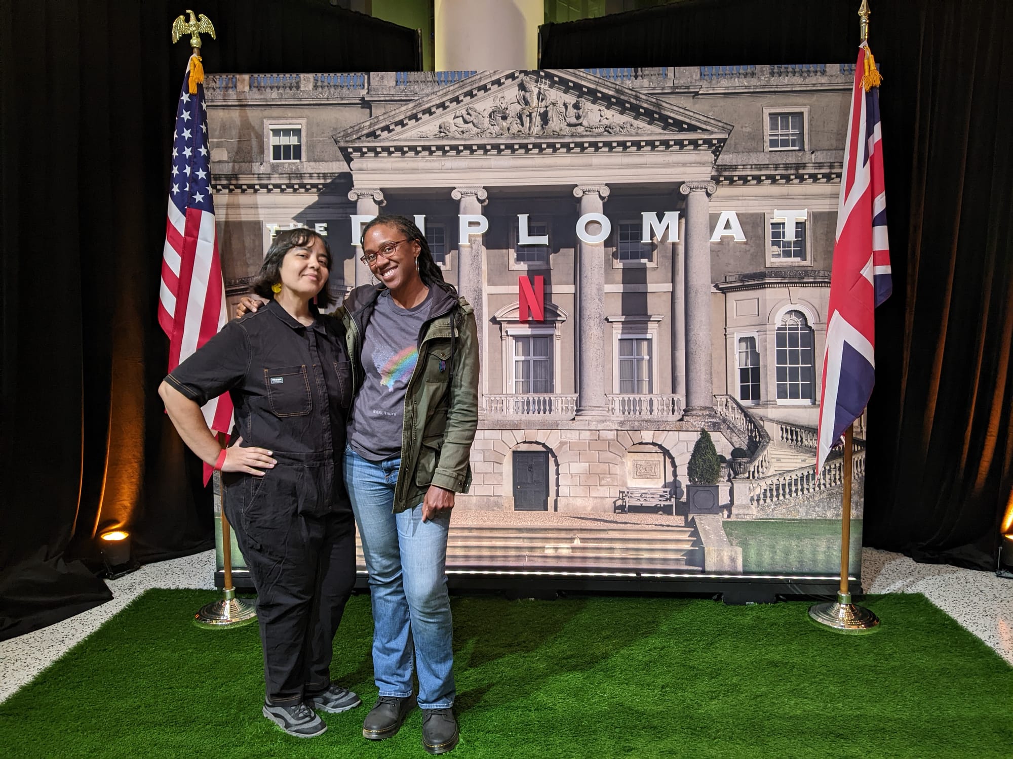 Image of a Latina in a black jumpsuit with a short, dark bob & a Black woman with long dark braids wearing jeans, a grey t-shirt, and a green jacket posing in front of a 'Diplomat' display.