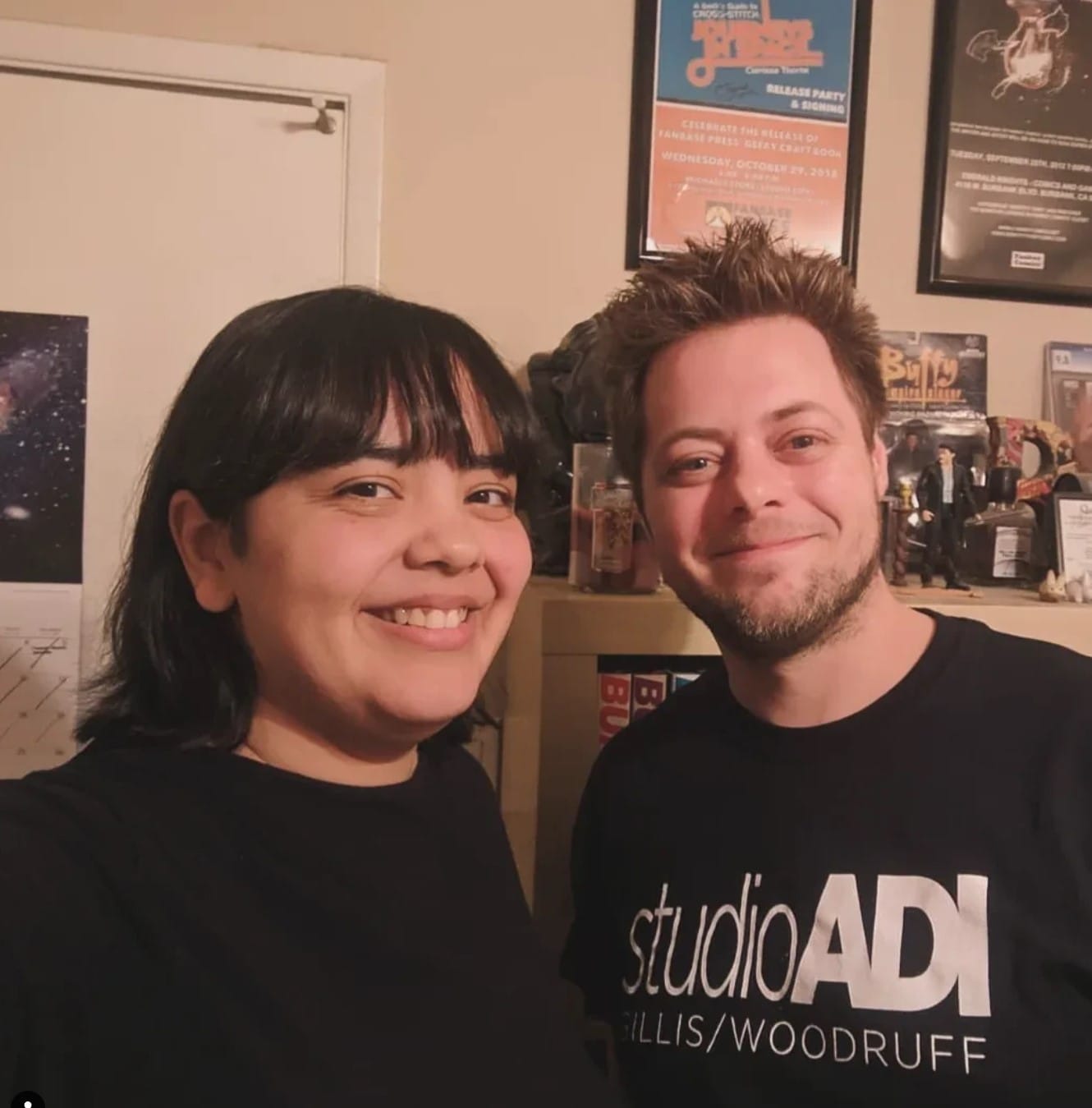 Teresa (brown Latina with a chin-length dark bob with bangs and wearing a black shirt) & Bryant (white man w/spiky brown hair and a beard wearing a black shirt) in a home office smiling.