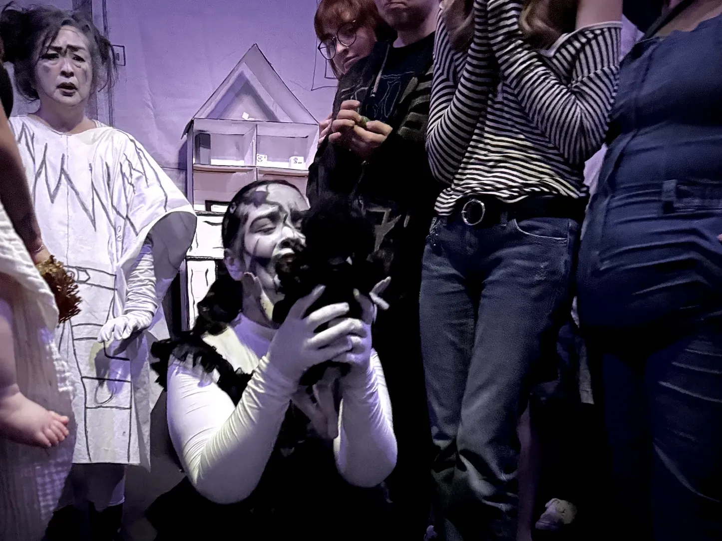 Image of a woman on her knees dressed all in black & white wearing black & white make-up crying as she looks at a doll while surrounded by onlookers.