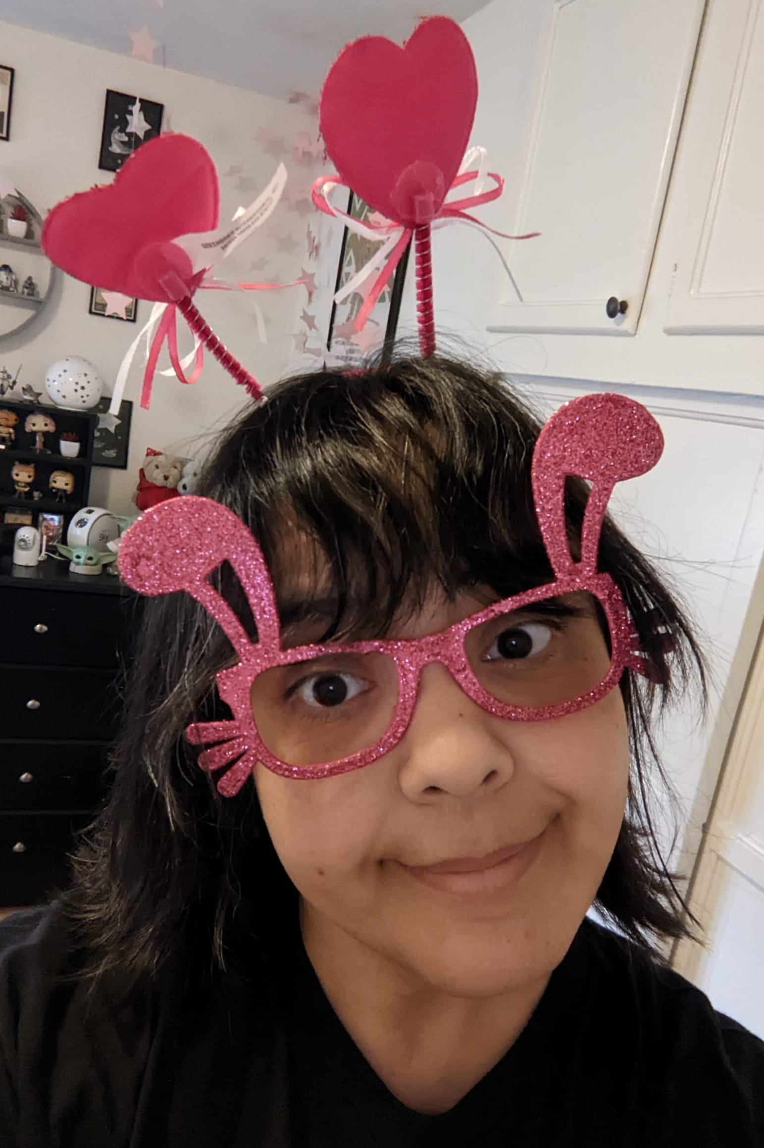 Image of Teresa Jusino (Latina with chin-length dark hair and bangs) wearing a black t-shirt, glittery pink glasses with bunny ears coming off them, and a headband with pink hearts sticking out of it.
