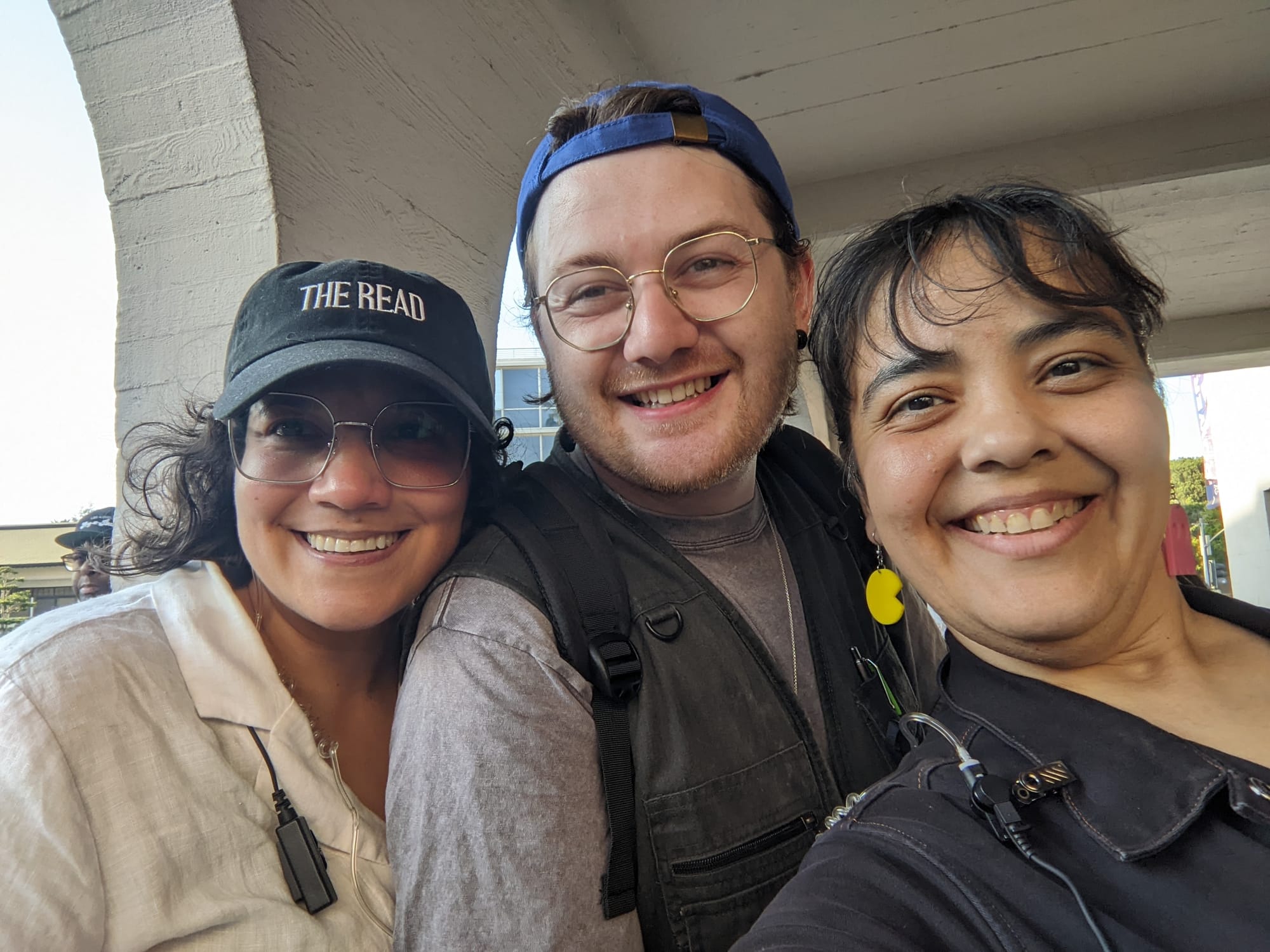 Color image of three people huddled for a selfie: a Latina with dark hair wearing a black baseball cap and glasses, a white man wearing a backwards blue baseball cap and glasses, and a Latina with dark hair and bangs wearing Pac-Man earrings.