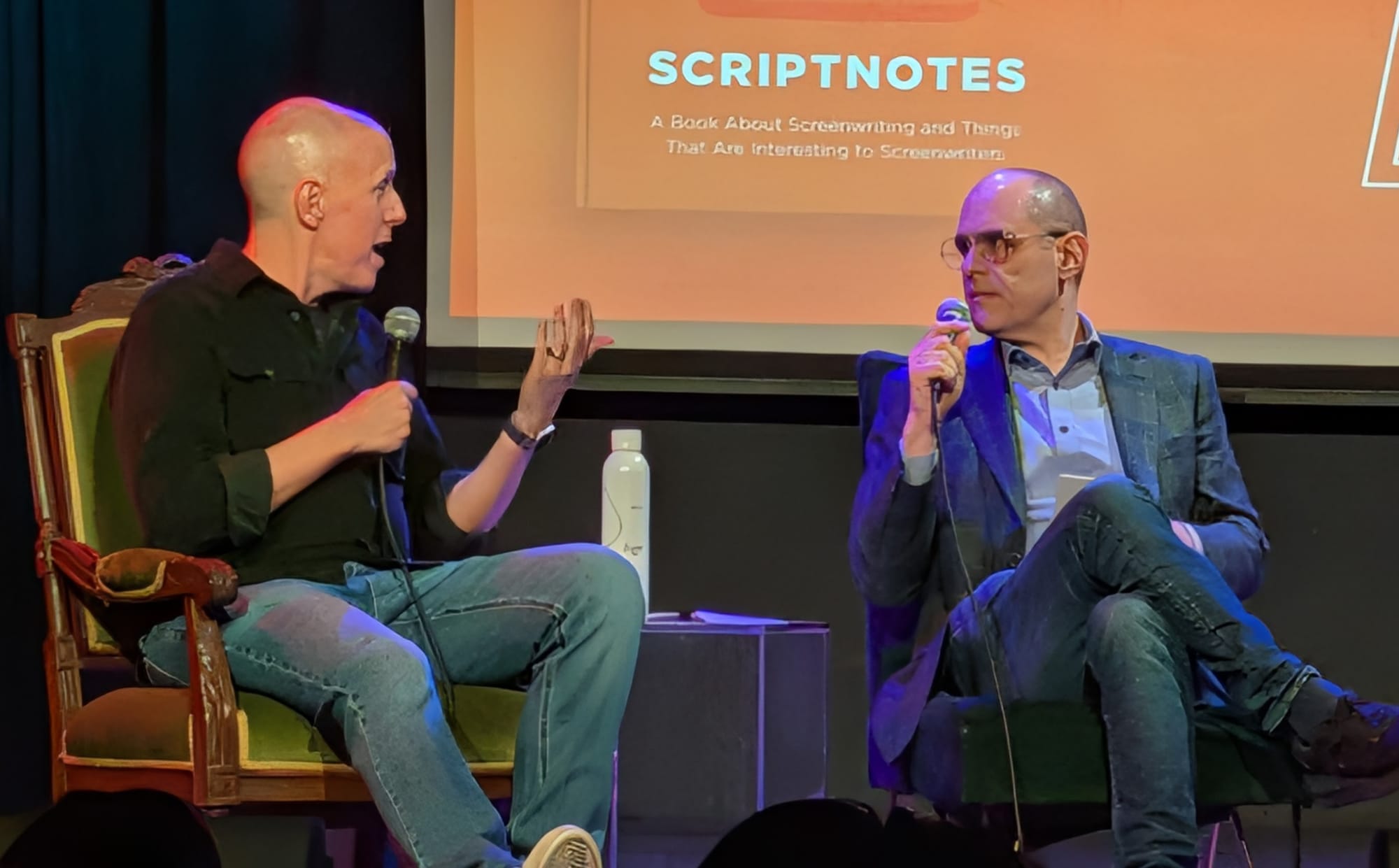 Image of John August and Craig Mazin sitting in chairs on a stage holding microphones at an event.