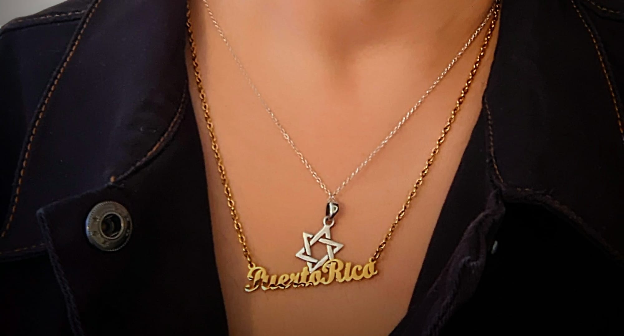 Image of 2 necklaces hanging from a woman's neck: one a silver Star of David, the other a gold nameplate that reads "Puerto Rico." They sit against the woman's bare brown skin visible through the top of an open, dark jumpsuit.