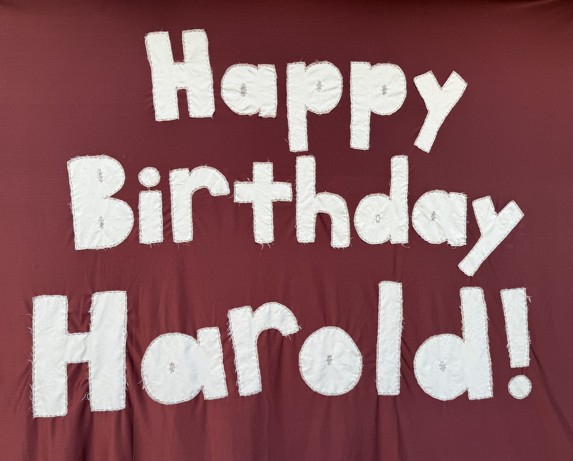 Image of brown banner made of fabric with white letters sewn on spelling out 'Happy Birthday Harold!' 