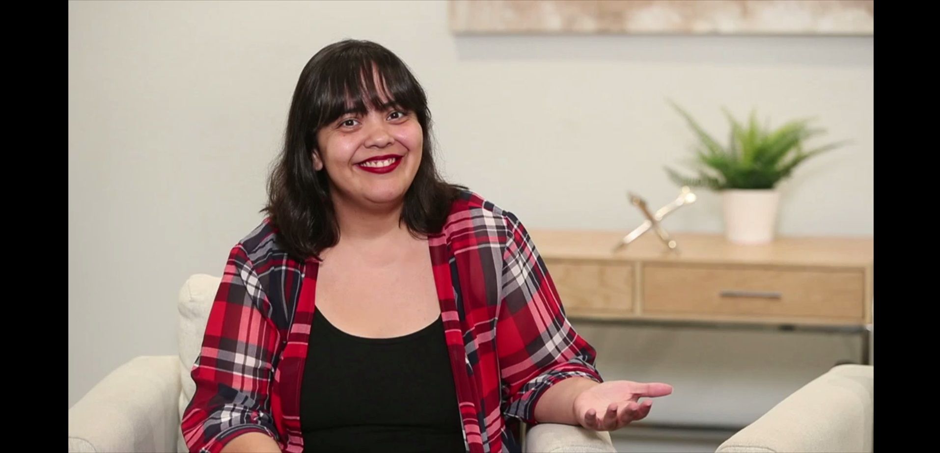 Teresa Jusino, olive-skinned woman w/shoulder-length dark hair & bangs sits in a studio.