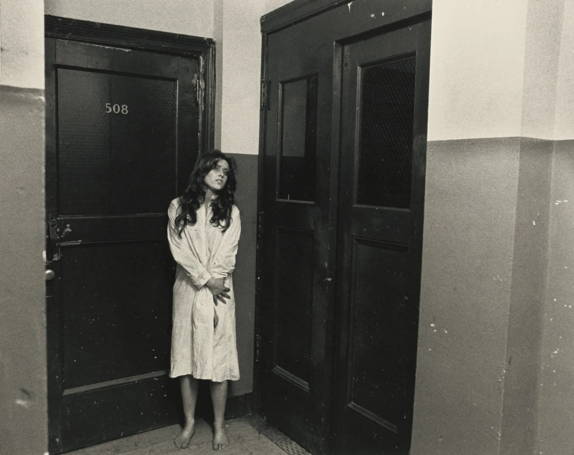 Black and white image of a white woman with long, dark hair standing in a hallway wearing a white bathrobe.