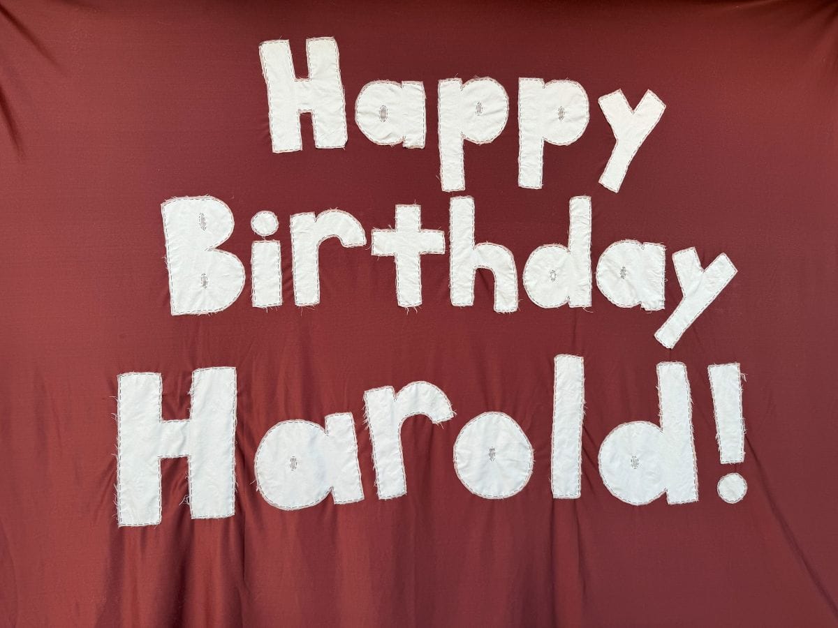 Close-up of a banner on brown fabric with white letters sewn on: it reads "Happy Birthday Harold!"