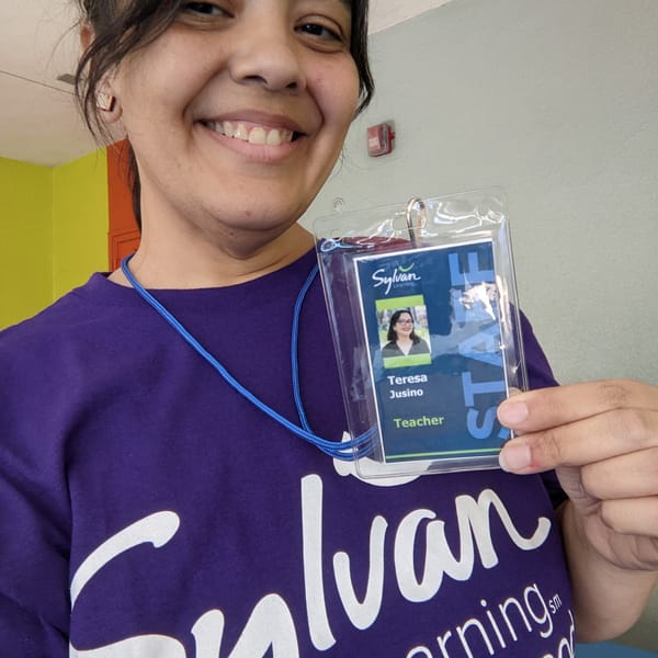 Image: brown Latina with dark hair in a ponytail smiling holding up a photo ID badge on a lanyard for Sylvan Learning Center.