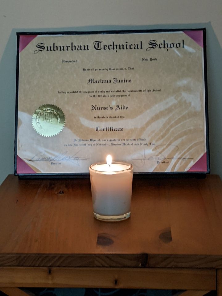 Image of a small, white, lit yahrzeit candle on a wooden tabletop in front of a graduation certificate.