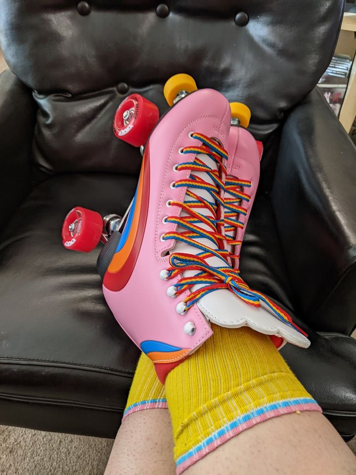 An image of feet crossed at the ankles wearing pink roller skates with rainbow designs on them and rainbow laces. 
